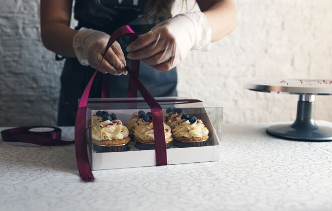 纸一个穿着灰色围裙的女孩在一个礼品盒里包上奶油纸杯蛋糕 把订单送到餐厅客户.烘焙在家里人盒子商店
