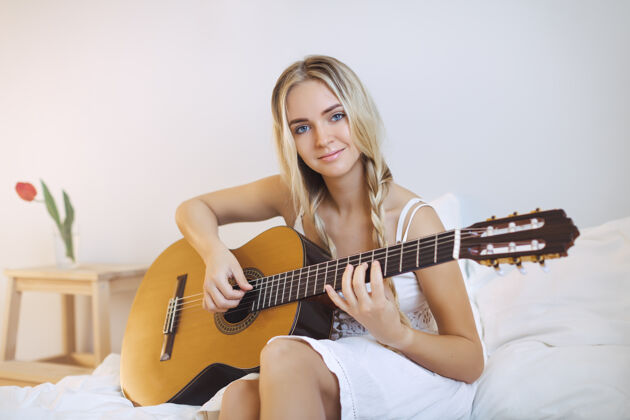生活方式美丽浪漫的年轻女子坐在床上弹吉他主页.肖像可爱的女孩活动乐器室内