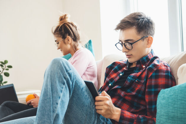 在线戴眼镜的白种人和他的妻子坐在沙发上 读着平板电脑和笔记本电脑上的东西公寓科技室内