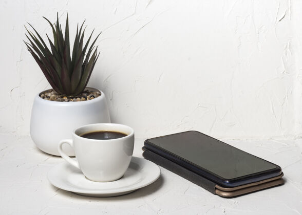 镜头白色抽象背景上带有黑色芳香咖啡的白色杯子 背景是一盆花 配智能手机爱早晨移动