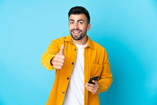 通讯高加索男子在孤立的蓝色背景下一边用手机一边竖起大拇指胡须手指西班牙裔