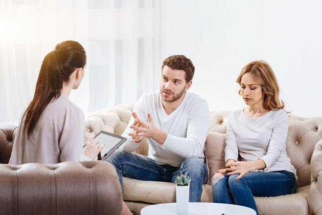 社交心理学session.帅哥一个不快乐的留胡子男人和他的妻子坐在沙发上 一边看着心理学家一边向她解释他们的问题预约解决方案咨询