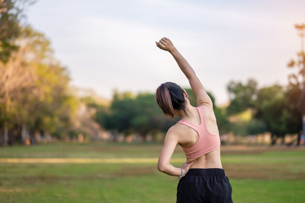 慢跑穿着粉色运动服的年轻成年女性在公园户外伸展肌肉 运动女性热身准备跑步和慢跑早上好健康 健身 运动和工作生活平衡概念锻炼女性健康
