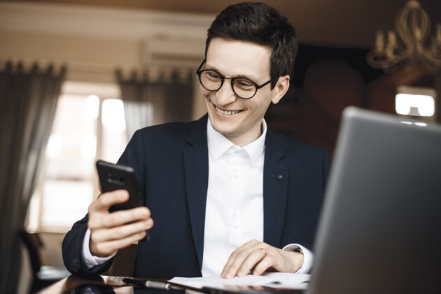 笔记本电脑一个年轻帅哥拿着智能手机 面带微笑地看着屏幕 在办公桌上的笔记本电脑前工作休闲应用程序活跃