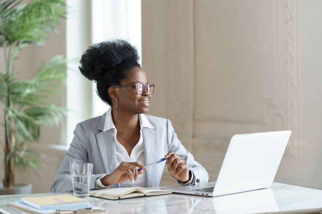 工作场所身着西装 面带微笑的非洲裔妇女戴着眼镜 在家里的办公室里用笔记本电脑工作 目光转向别处美国工作站放松