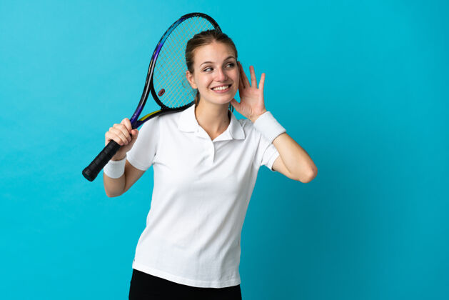 健康年轻的女子网球运动员被隔离在蓝色背景上 用手捂着耳朵听东西交流玩活动