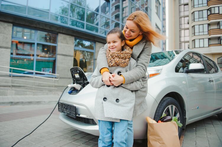 充电妈妈带着女儿在电动加油站给电动车充电 用手机说话生活方式当代移动性