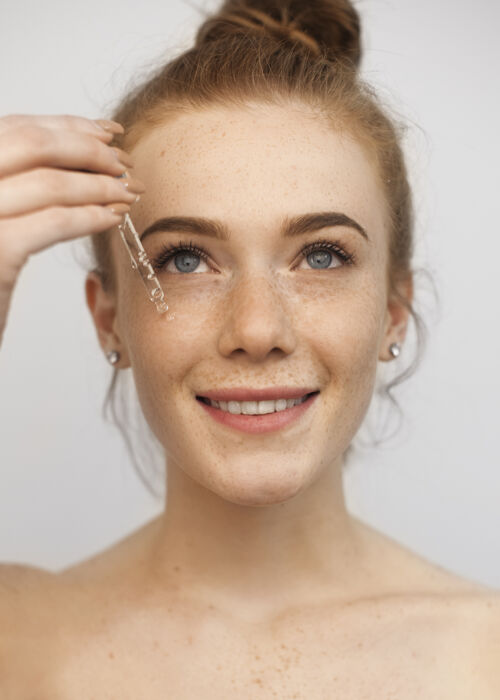 美丽一个年轻漂亮的红发女人用滴管涂透明质酸的特写照片 微笑着隔离在白墙上化妆品保湿乳液
