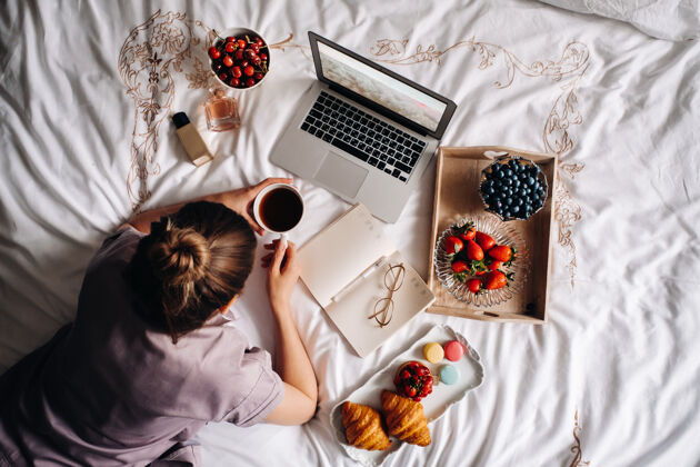 工作一个女孩晚上坐在床上 手里拿着智能手机 吃着草莓 一个女孩睡觉前吃着糖果舒适互联网室内