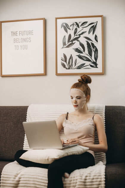 聊天可爱的年轻女性影响者坐在家里的沙发上 一边用笔记本电脑工作 一边戴着护肤面膜放松室内室内