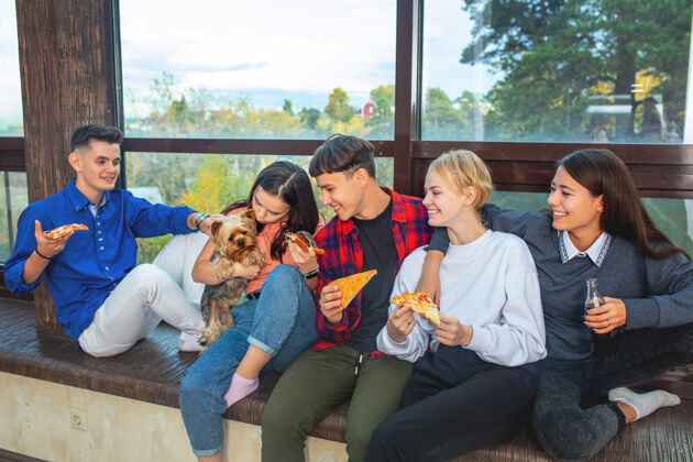 快一群年轻快乐的朋友带着宠物狗坐在家里的窗户上吃比萨饼女食物人
