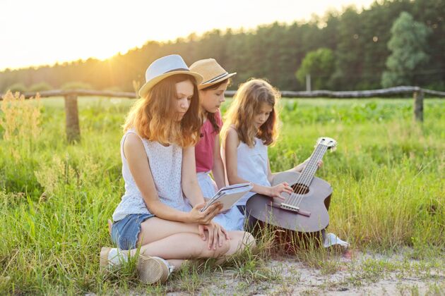 草带古典吉他的女孩自然孩子们在草坪上放松 学习弹吉他 唱歌 夏日草地背景上的日落孩子青少年户外