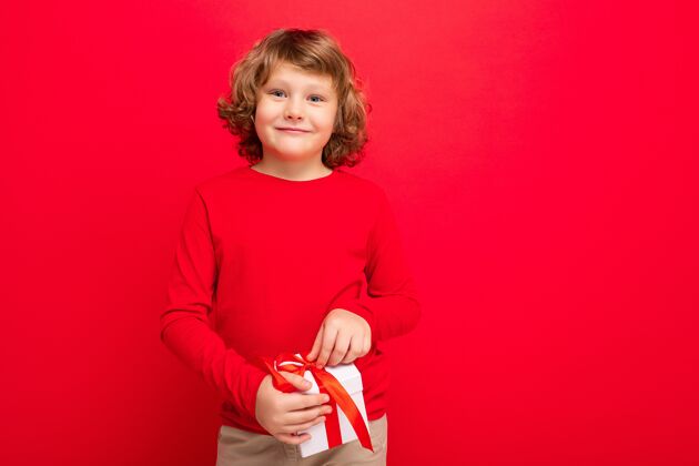 童年正面微笑的金发卷发男孩隔着红色背景墙穿着红色毛衣拿着礼盒孩子帅气快乐