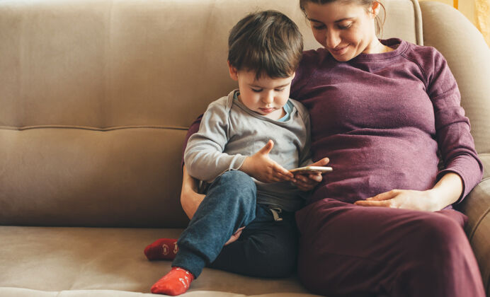 沙发满脸雀斑的高加索孕妇坐在沙发上 她的小男孩在用电话产前孕妇电话