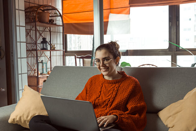 年轻人一个戴着眼镜的年轻女子在沙发上对着相机微笑 一边用笔记本电脑工作和学习青年个人电子
