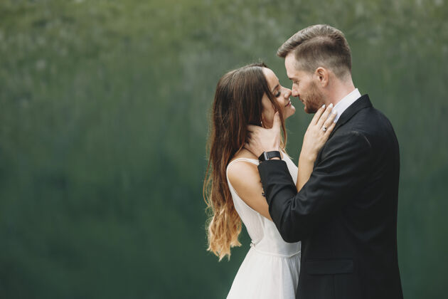 新郎一对美丽的已婚夫妇在绿色的湖面上亲吻前互相对视的侧视图浪漫自然订婚