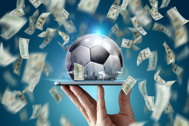使用在线体育下注美元一只手拿着一部智能手机和一个足球球创意背景 赌博应用程序球目标