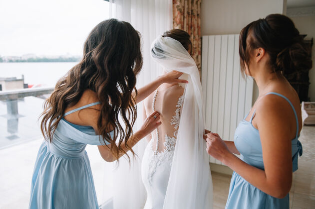 早晨伴娘在婚礼前帮新娘系上白色婚纱的花边优雅礼服新娘