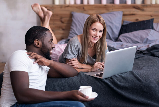 喜悦甜蜜的泰晤士报很高兴一对年轻的国际情侣在卧室里 一边用笔记本电脑 一边微笑关系放松服务