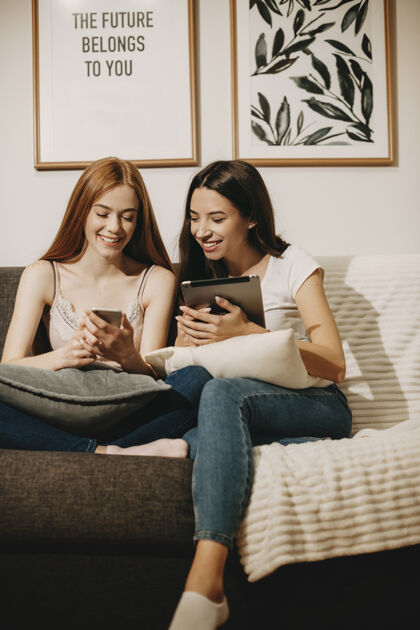 学生两个可爱的年轻女性坐在沙发上看着他们的智能手机笑全长微笑雀斑女人
