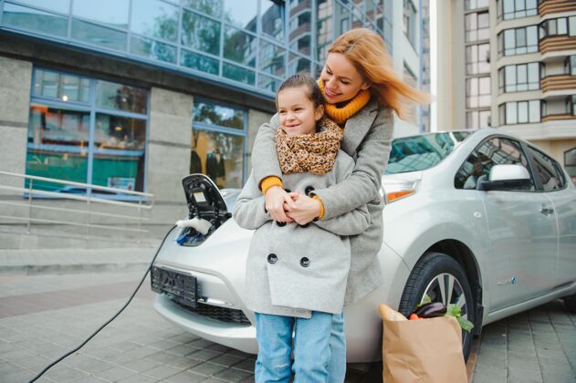 充电妈妈带着女儿在电动加油站给电动车充电 用手机说话生活方式当代移动性