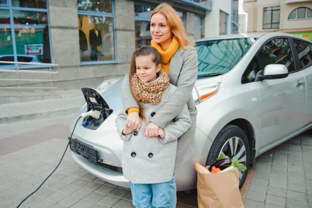 工业妈妈带着女儿在电动加油站给电动车充电 用手机说话电子替代儿童