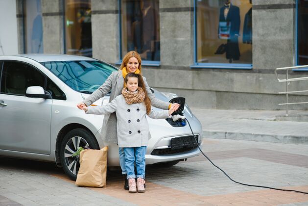 汽车妈妈带着女儿在电动加油站给电动车充电 用手机说话电力汽车替代