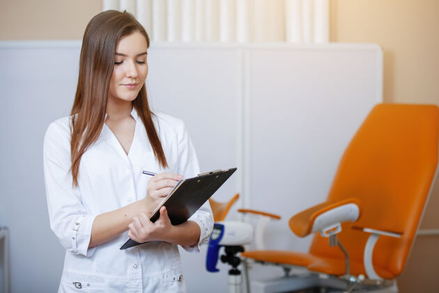 诊所实习医生年轻漂亮的女人 穿着白衣服 手里拿着一块药片 在橙色的妇科手术椅旁对着镜头摆姿势医院医疗健康