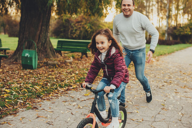 成人可爱的小女孩骑着自行车 而她的父亲正试图抓住她在公园的户外笑户外自行车秋天