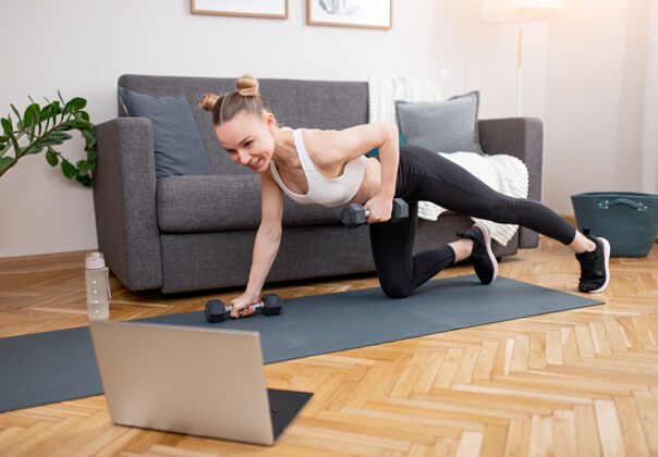 努力适合年轻的女运动员做哑铃锻炼和看视频在笔记本电脑上在线锻炼在家里垫子运动服视频