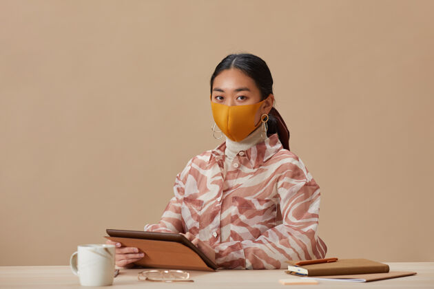 职业戴着防护面具的亚洲女性坐在桌旁使用数码平板电脑时注视前方的肖像防护面具技术学生