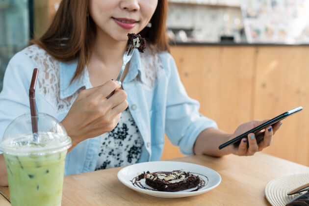 女性亚洲女人一边用智能手机一边吃蛋糕玻璃新鲜美味