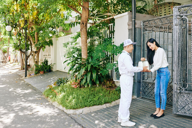微笑一个穿着白色制服的送奶工正在给一个年轻漂亮的亚洲女人送一套玻璃瓶装牛奶盒子站立女人