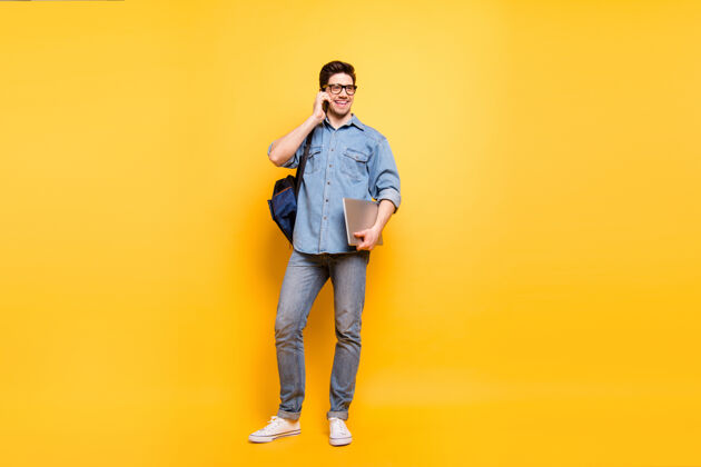 通话一张全身尺寸的照片 照片上是一个快乐的 积极的 满嘴笑容的男人 手里拿着笔记本电脑 穿着白色运动鞋打电话 隔着一堵充满活力的彩色墙学习交谈背包