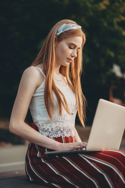 雀斑一位红发雀斑美女坐在公园的长凳上看着笔记本电脑屏幕的侧视图公园打字年轻