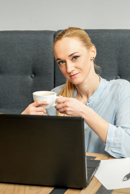 一位年轻的女商人坐在咖啡馆的桌子旁 手里拿着笔记本电脑 手里拿着一杯咖啡自由职业者商店计算机