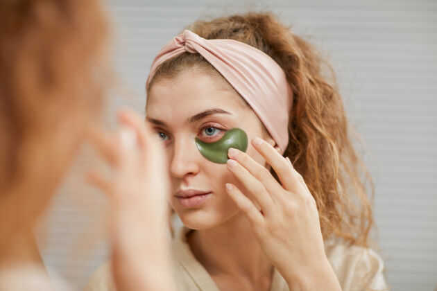 欢呼年轻的女人在照镜子的时候 在眼睛下面敷上眼罩肖像化妆贴片