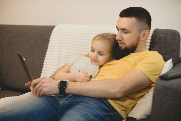 父母一位留着胡须的年轻父亲的侧视图 他和女儿在家里靠在沙发上看平板电脑自由空间年轻人教育