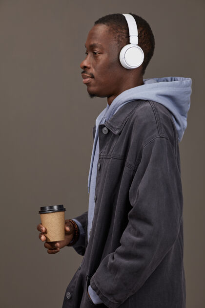 休闲装侧视图：穿着休闲服的年轻人戴着耳机听音乐 喝着咖啡 孤零零地站在灰色的墙上肖像成人站着