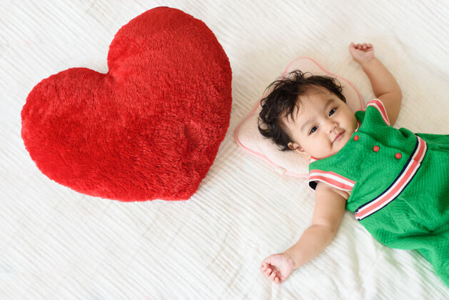 爱一个可爱的小女婴穿着绿色的裙子躺在床上一个红色的心形枕头旁边婴儿天真甜蜜