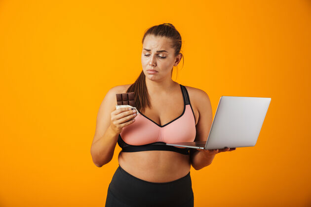 健身一个穿着运动服的失望的超重年轻女子的肖像 孤立地站在黄色的墙上 用笔记本电脑 拿着巧克力棒瑜伽损失白种人