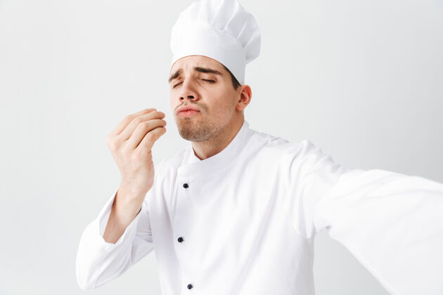 外套穿着制服的快乐厨师长孤立地站在白墙上 自拍帽子美食家专业