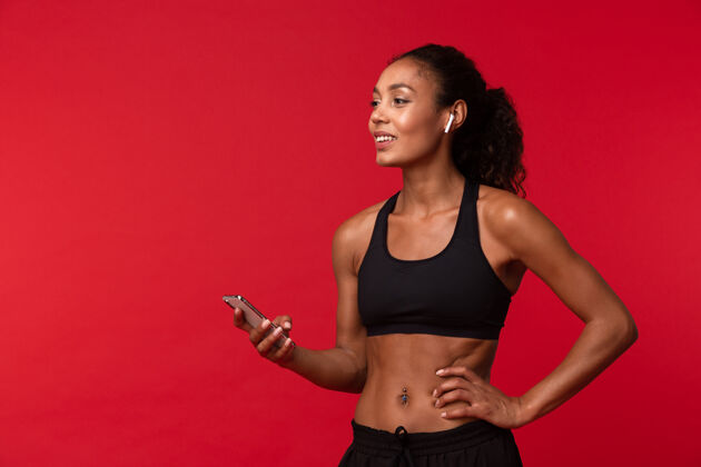 运动装一个穿着黑色运动服 使用智能手机和无线耳机 隔着红墙的美丽的非洲裔美国女人的肖像运动运动服20多岁