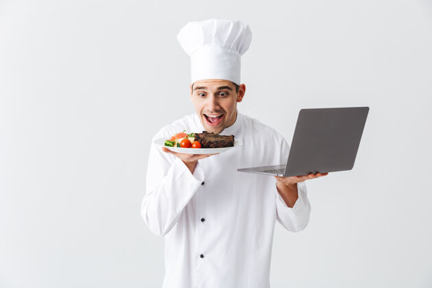 帅气兴奋的厨师穿着制服站在白色的墙上 拿着笔记本电脑 展示着一道菜制服笔记本电脑沟通