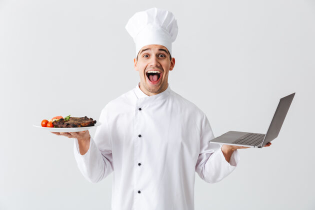 盘子兴奋的厨师穿着制服站在白色的墙上 拿着笔记本电脑 展示着一道菜菜肴互联网帽子