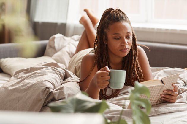 房子年轻的女人躺在床上喝咖啡 早上看书 她享受她的周末放松休息舒适