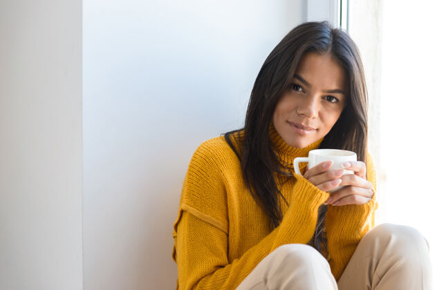 冬天一个穿着毛衣的可爱的年轻女子坐在室内的窗边 端着一杯茶的特写镜头季节茶微笑