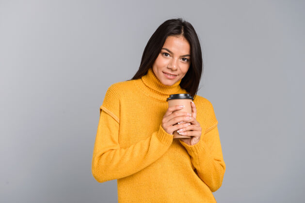看起来一个微笑的年轻女子穿着毛衣 拿着外卖咖啡杯 隔着灰色的墙吸引力茶手臂
