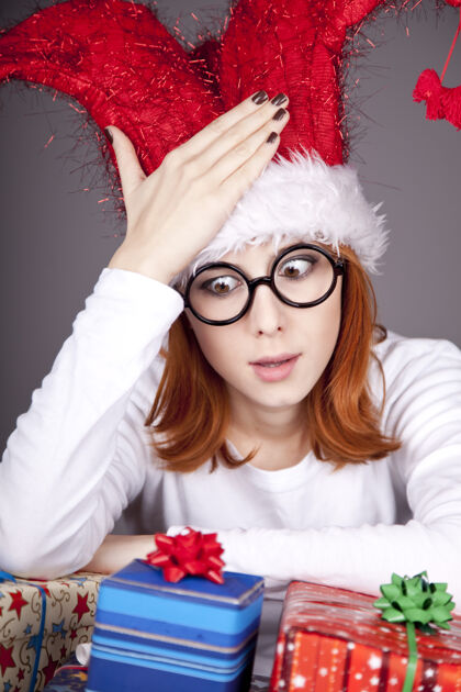 服装滑稽的红发女孩戴着圣诞帽 戴着带礼盒的眼镜盒子长礼物