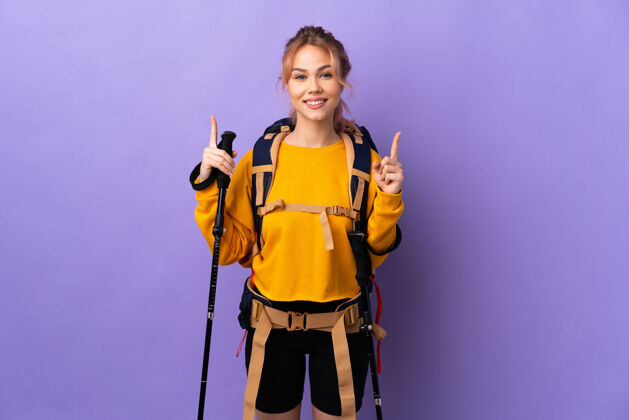 休闲女孩背着背包和登山竿在孤立的紫色上指点着一个好主意眼睛手指小径